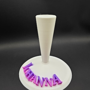 Puede incluir: Un soporte para pastel de pl&aacute;stico blanco con un poste central c&oacute;nico y alto. La base del soporte tiene el nombre "Johanna" escrito en letras rosas y moradas.
