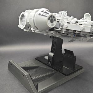 以下が含まれることがあります： 黒いディスプレイスタンドに置かれたグレーとブラックのLEGOミレニアムファルコンモデル。スタンドはテクスチャ加工された表面と、モデルを置くための盛り上がったプラットフォームを持っています。