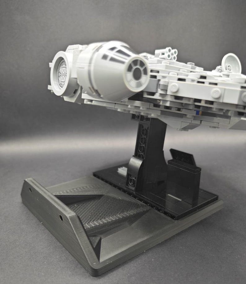 以下が含まれることがあります： 黒いディスプレイスタンドに置かれたグレーとブラックのLEGOミレニアムファルコンモデル。スタンドはテクスチャ加工された表面と、モデルを置くための盛り上がったプラットフォームを持っています。