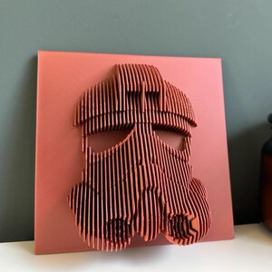 Puede incluir: Una pieza de arte de pared de madera roja que representa una cara estilizada con un casco. El casco est&aacute; hecho de muchas tiras de madera delgadas y verticales.