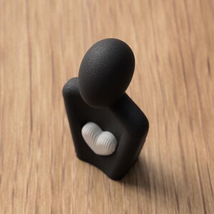 May include: A black abstract sculpture with a textured head and body, featuring a white heart-shaped detail. The sculpture is set against a wooden background, creating a contrast between the smooth and rough textures.