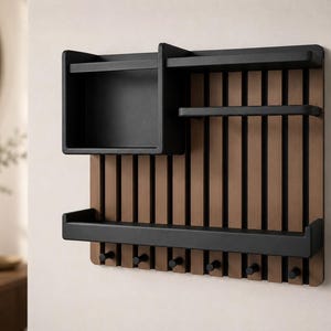 May include: A wall-mounted organizer featuring a slatted brown wood back panel with a black frame. It includes a square storage box, two horizontal shelves, and six small black hooks along the bottom edge for hanging various items.