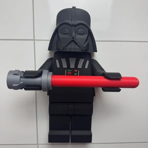May include: A black and grey Lego figurine of Darth Vader holding a red lightsaber. The figurine is mounted on a white tiled wall.