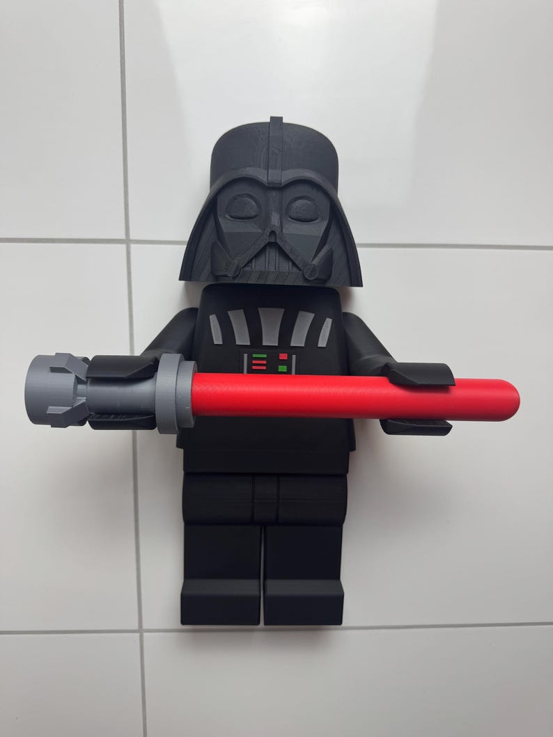 May include: A black and grey Lego figurine of Darth Vader holding a red lightsaber. The figurine is mounted on a white tiled wall.