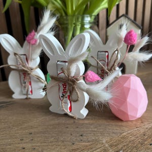 May include: Three white ceramic bunny figurines with brown twine and pink flowers. Each bunny holds a small chocolate bar with the word "Kinder" printed on the wrapper.