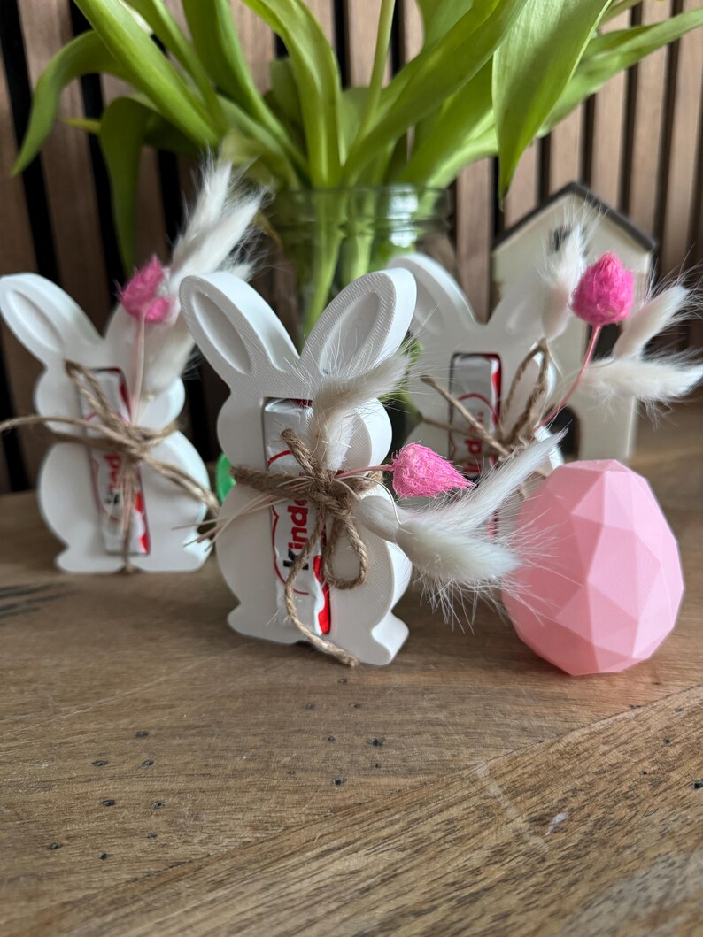 May include: Three white ceramic bunny figurines with brown twine and pink flowers. Each bunny holds a small chocolate bar with the word "Kinder" printed on the wrapper.