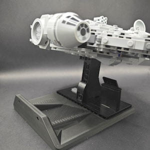 以下が含まれることがあります： 黒いディスプレイスタンドに置かれた、グレーとブラックのLEGOミレニアム・ファルコンの宇宙船モデル。宇宙船は横に傾いており、モデルの下側が見えるようになっている。
