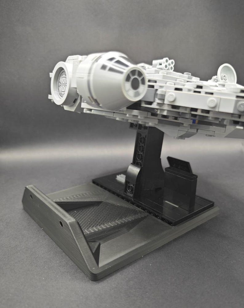 以下が含まれることがあります： 黒いディスプレイスタンドに置かれた、グレーとブラックのLEGOミレニアム・ファルコンの宇宙船モデル。宇宙船は横に傾いており、モデルの下側が見えるようになっている。