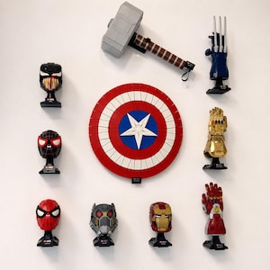 Puede incluir: Una colecci&oacute;n de sets LEGO con tem&aacute;tica de superh&eacute;roes exhibidos en una pared blanca. El conjunto incluye el escudo del Capit&aacute;n Am&eacute;rica, el martillo de Thor y varios bustos de personajes como Venom, Spider-Man e Iron Man. La paleta de colores est&aacute; dominada por el rojo, el azul, el dorado y el gris.