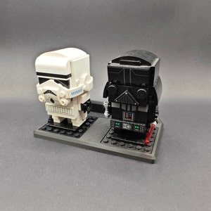 Może przedstawiać: Zestaw minifigurek Lego w kolorze czarnym i białym przedstawiający Dartha Vadera i szturmowca. Minifigurki są umieszczone na czarnej podstawie z czerwonym mieczem świetlnym.