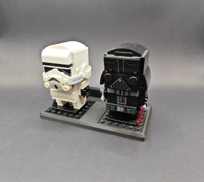 Może przedstawiać: Zestaw minifigurek Lego w kolorze czarnym i białym przedstawiający Dartha Vadera i szturmowca. Minifigurki są umieszczone na czarnej podstawie z czerwonym mieczem świetlnym.