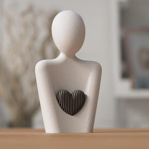 May include: A textured, off-white sculpture of a person holding a heart. The heart is made of dark, vertical wooden slats. The sculpture has a minimalist design and is placed on a wooden surface.
