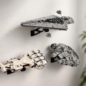 May include: Three Star Wars Lego starships mounted on a white wall. The ships are grey and white, with black mounting brackets. The top ship is a large grey model, while the other two are smaller and white.