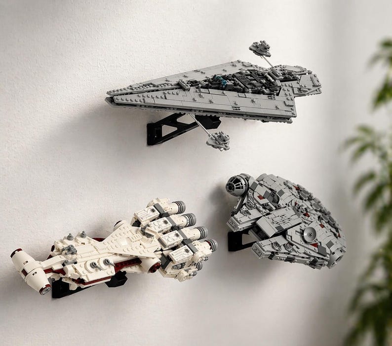 May include: Three Star Wars Lego starships mounted on a white wall. The ships are grey and white, with black mounting brackets. The top ship is a large grey model, while the other two are smaller and white.