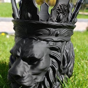 May include: A black lion head planter with a crown. The planter is made of a dark material and features detailed carvings of the lion's mane, face, and crown. The crown has decorative elements and is set against a backdrop of green grass.