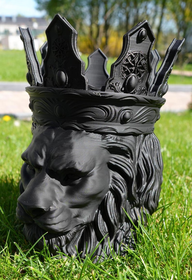 May include: A black lion head planter with a crown. The planter is made of a dark material and features detailed carvings of the lion's mane, face, and crown. The crown has decorative elements and is set against a backdrop of green grass.