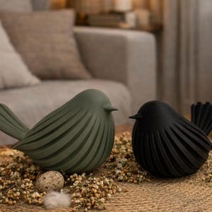 May include: Two decorative bird sculptures, one olive green and one black, are displayed on a round woven mat with dried flowers and an egg. The birds have a stylized design with ribbed wings and bodies. A lit candle and a decorative lantern are in the background.