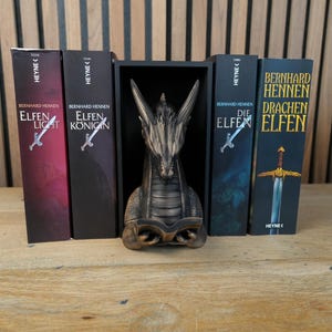 May include: A collection of fantasy books with titles like "Elfen Light" and "Elfen Konigin" by Bernhard Hennen, displayed with a dragon statue. The books have a sword illustration. The dragon statue is bronze-colored.