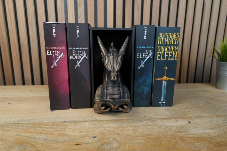 May include: A collection of fantasy books with titles like "Elfen Light" and "Elfen Konigin" by Bernhard Hennen, displayed with a dragon statue. The books have a sword illustration. The dragon statue is bronze-colored.