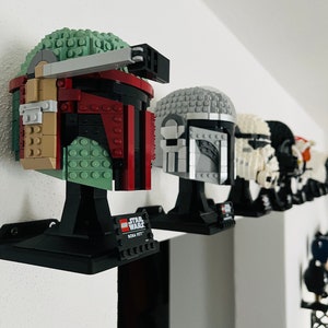 Könnte beinhalten: Eine Sammlung von fünf LEGO Star Wars Helmmodellen, die auf schwarzen Ständern montiert sind. Die Helme sind Boba Fett, Darth Vader, Stormtrooper, TIE-Jäger-Pilot und ein Mandalorianer. Die Helme sind alle in verschiedenen Farben gehalten und haben unterschiedliche Details. Die Ständer tragen das LEGO Star Wars Logo und den Namen des Charakters.