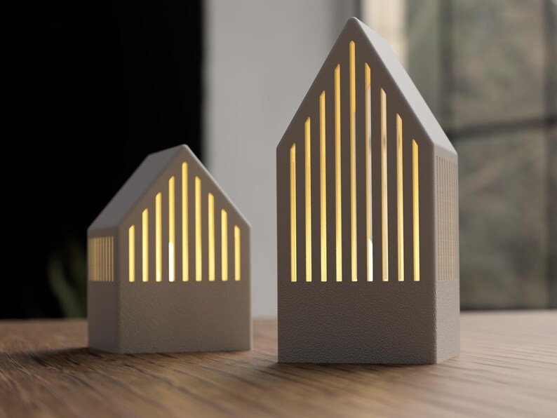 May include: Two off-white, house-shaped decorative lamps. Each lamp has a gabled roof and vertical cutouts that emit a warm, yellow light. The lamps are placed on a wooden surface.