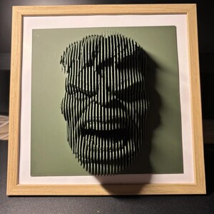 May include: A framed 3D art piece of a face made of stacked wood strips. The face is green and has a smiling expression. The frame is made of light wood.