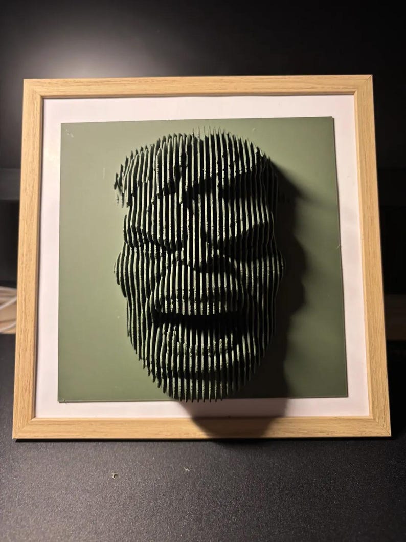 May include: A framed 3D art piece of a face made of stacked wood strips. The face is green and has a smiling expression. The frame is made of light wood.