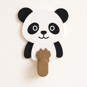 May include: A wall-mounted hook shaped like a panda face with black and white features. The panda has black ears, eye patches, and a nose. A brown, textured paw print design is positioned on the panda's chest, extending into a vertical hook.