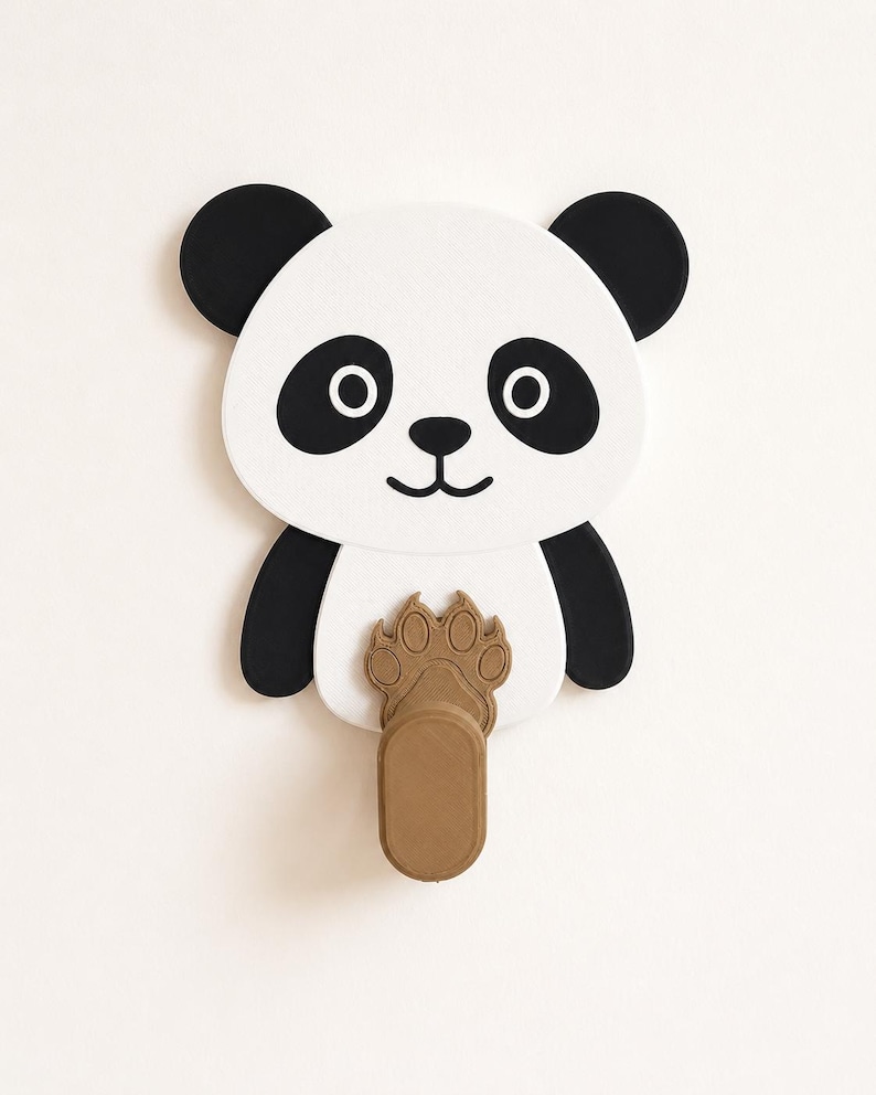 May include: A wall-mounted hook shaped like a panda face with black and white features. The panda has black ears, eye patches, and a nose. A brown, textured paw print design is positioned on the panda's chest, extending into a vertical hook.