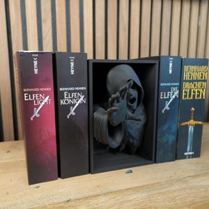 May include: A selection of German fantasy novels by Bernhard Hennen, including "Elfen Licht" and "Elfen K&ouml;nigin", with a dark figure statue in a black display case. The books feature a sword graphic. A small potted plant is also present.