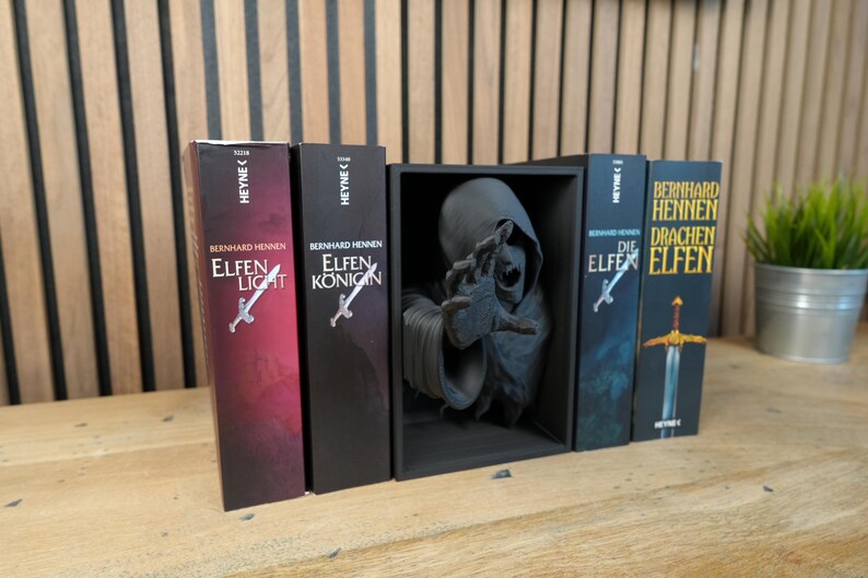 May include: A selection of German fantasy novels by Bernhard Hennen, including "Elfen Licht" and "Elfen K&ouml;nigin", with a dark figure statue in a black display case. The books feature a sword graphic. A small potted plant is also present.