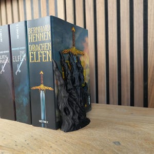 K&ouml;nnte beinhalten: Eine Reihe von Fantasy-Romanen mit einem schwarzen, schwertf&ouml;rmigen Buchst&uuml;tzen. Die Buchst&uuml;tze ist dunkel und hat ein detailliertes Design. Die Buchcover sind in verschiedenen Farben gehalten, mit Titeln auf Deutsch. Die B&uuml;cher stehen auf einer Holzoberfl&auml;che.