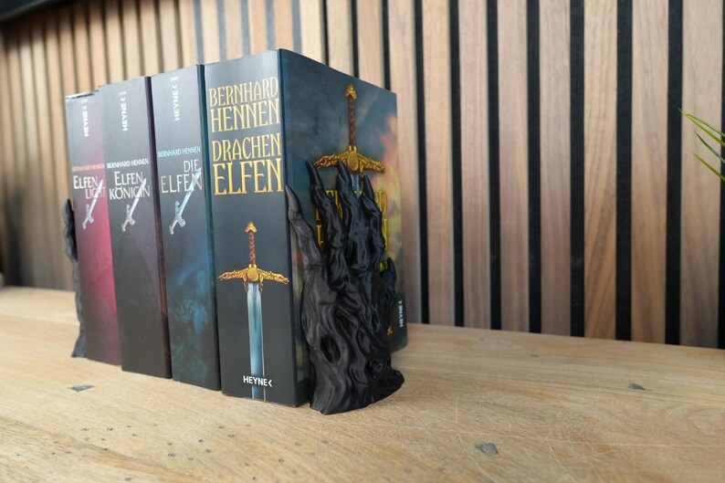 K&ouml;nnte beinhalten: Eine Reihe von Fantasy-Romanen mit einem schwarzen, schwertf&ouml;rmigen Buchst&uuml;tzen. Die Buchst&uuml;tze ist dunkel und hat ein detailliertes Design. Die Buchcover sind in verschiedenen Farben gehalten, mit Titeln auf Deutsch. Die B&uuml;cher stehen auf einer Holzoberfl&auml;che.