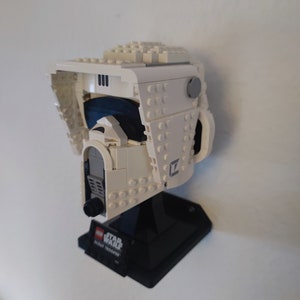 Puede incluir: Casco de Scout Trooper Lego Star Wars blanco con una base negra. El casco tiene una visera azul y un detalle gris en el lateral. La base tiene las palabras "LEGO STAR WARS SCOUT TROOPER" impresas.