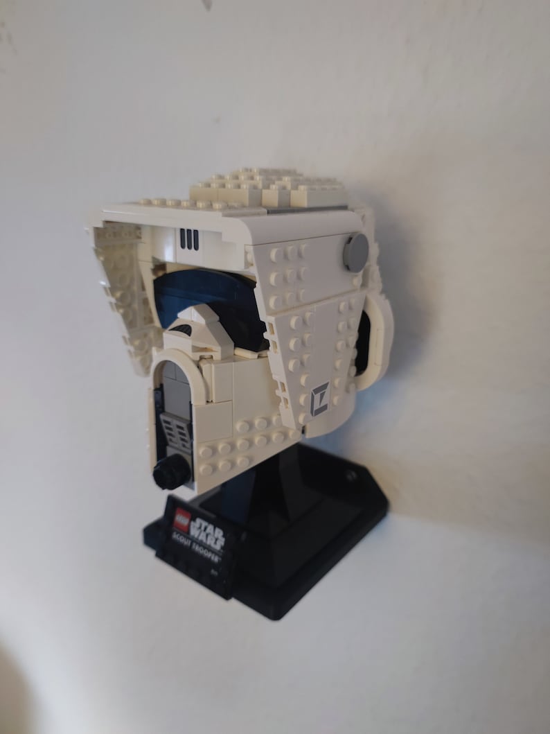 Puede incluir: Casco de Scout Trooper Lego Star Wars blanco con una base negra. El casco tiene una visera azul y un detalle gris en el lateral. La base tiene las palabras "LEGO STAR WARS SCOUT TROOPER" impresas.