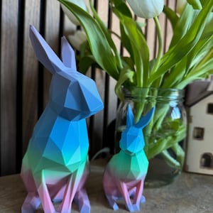 May include: Two geometric rabbit figurines, one larger than the other, are made of a gradient of blue, green, and pink plastic. The figurines are sitting on a wooden surface in front of a vase of white tulips.