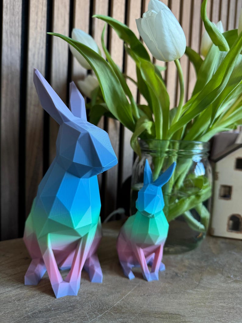 May include: Two geometric rabbit figurines, one larger than the other, are made of a gradient of blue, green, and pink plastic. The figurines are sitting on a wooden surface in front of a vase of white tulips.