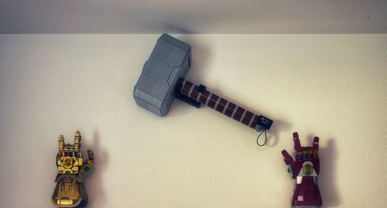 Op de afbeelding: Een display met Lego-modellen: Thor's hamer, Mjolnir, en twee Infinity Gauntlets. De hamer is grijs en bruin, de handschoenen zijn goud en rood. De items zijn op een witte muur gemonteerd, wat een superheldenthema cre&euml;ert.