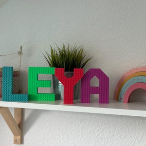 May include: A shelf displays the name "LEYA" spelled out with colorful building block letters. The letters are teal, green, red, and purple. A small plant in a metal pot and a wooden rainbow are also on the shelf.