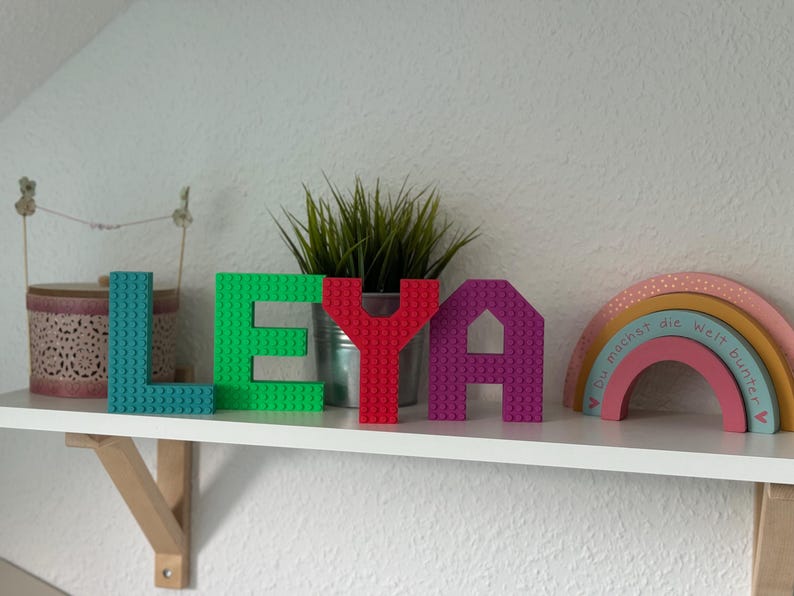 May include: A shelf displays the name "LEYA" spelled out with colorful building block letters. The letters are teal, green, red, and purple. A small plant in a metal pot and a wooden rainbow are also on the shelf.