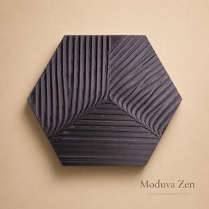 May include: A dark gray, hexagonal wall art piece with a textured, carved design. The surface is divided into sections with vertical and diagonal grooves, creating a leaf-like pattern. The text "Moduva Zen" is at the bottom.