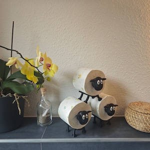May include: Three black sheep-shaped toilet paper holders stacked on top of each other. The holders are made of plastic and have a white sheep design. The holders are on a black surface.