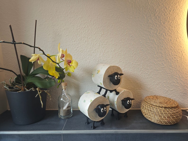 May include: Three black sheep-shaped toilet paper holders stacked on top of each other. The holders are made of plastic and have a white sheep design. The holders are on a black surface.