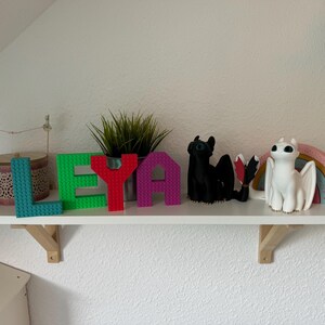 May include: A white shelf displays colorful decorative items. The word "LEYA" is spelled out with teal, green, red, and purple block letters. A black dragon figurine and a white dragon figurine with a rainbow are also on the shelf.