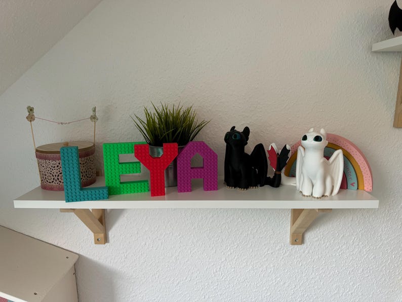 May include: A white shelf displays colorful decorative items. The word "LEYA" is spelled out with teal, green, red, and purple block letters. A black dragon figurine and a white dragon figurine with a rainbow are also on the shelf.