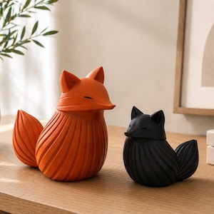 K&ouml;nnte beinhalten: Zwei dekorative Fuchsstatuen, eine orangefarbene und eine schwarze. Beide haben ein stilisiertes Design mit einem strukturierten, geriffelten K&ouml;rper und einem spitzen Schwanz. Die F&uuml;chse stehen auf einer Holzoberfl&auml;che, passend f&uuml;r die Inneneinrichtung.