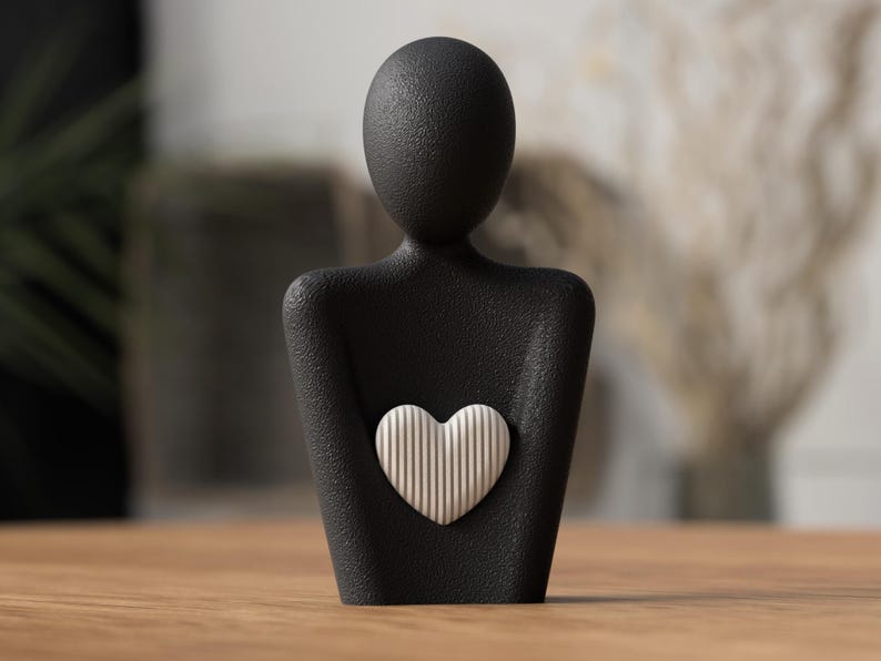 May include: A black sculpture of a person holding a white heart with vertical lines. The sculpture has a textured surface and is set on a wooden table. The background is blurred, with hints of greenery and neutral tones.