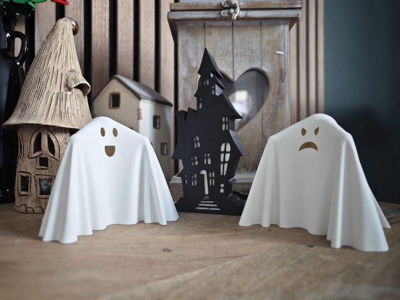 May include: Two white ghost figurines with different facial expressions. The ghost on the left is smiling, and the ghost on the right is frowning. The ghosts are sitting on a wooden surface in front of a black silhouette of a haunted house.