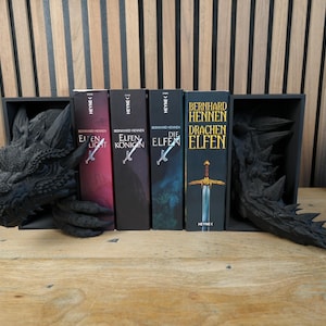 May include: A black dragon-themed bookshelf with four fantasy novels. The dragon head and tail bookends frame the books, which have titles in German. A small potted plant sits to the right.