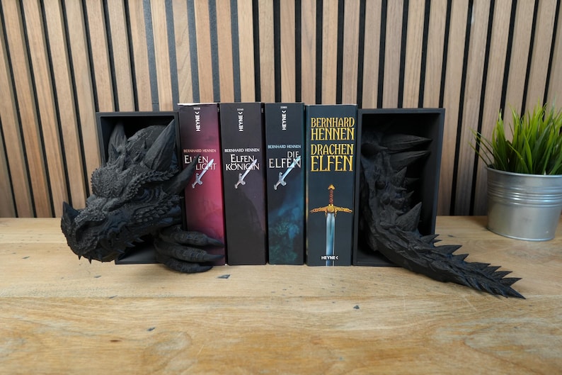 May include: A black dragon-themed bookshelf with four fantasy novels. The dragon head and tail bookends frame the books, which have titles in German. A small potted plant sits to the right.